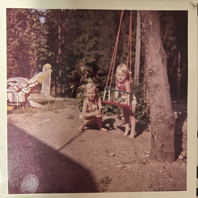 Kaksi pientä tyttöä omakotitalon pihalla, kameraan katsoen. Toinen nojaa puusta ripustettuun pikkulasten keinuun, toinen istuu kyykyssä.