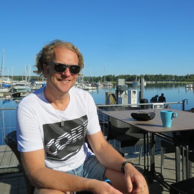 En man med blont lockigt hår, solglasögon, t-skjorta och shorts sitter på en uteservering vid en hamn. (Ekenäs gästhamn)