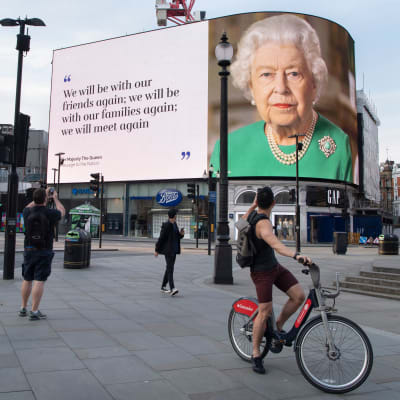 Fotgängare och en cyklist tittar upp mot den stora skärmen vid Piccadilly Circus som pryds av drottning Elizabeths porträtt och citatet "We will be with our friends again; we will be with our families again; we will meet again.  