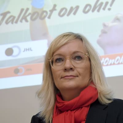 JHL:s ordförande Päivi Niemi-Laine tittar snett in i kameran. Bakom henne finns en bild av en städare på ett sjukhus med texten "Talkoot on ohi" alltså "Talkot är slut".