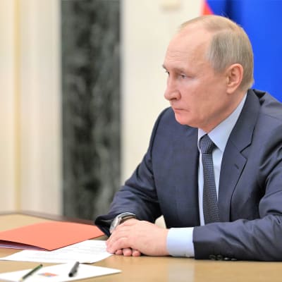 Putin istuu pöydän ääressä tummanharmaassa puvussa, vaaleansinisessä kauluspaidassa, tumma kravatti kaulassaan. Pöydällä edessä on pari paperia ja kynää ja oranssi kansio. Putinin ilme on vakava.