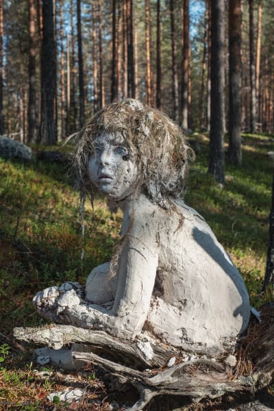 Lapsenmuotoinen vaaleasta massasta muotoiltu patsas, hiukset sammalta, katsoo kuvasta vasemmalle, taustalla metsää