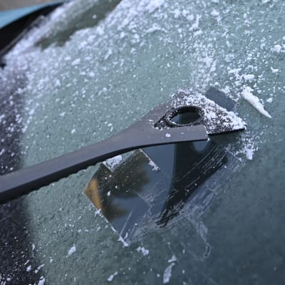 En person skrapar bort is från en bils vindruta. Det enda som syns är en hand, en skrapa och is på vindrutan.