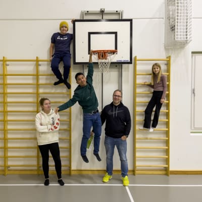 Fyra barn och en vuxen står i en gymnastisal. Tre av barnen har klättrat uppför ribbor som finns vid väggen, en av dem hänger till och med i en basketkorg.