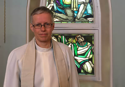 Prästen Rose-Maj Friman klädd i vit alba står framför fönster med glaskonst i Brändö kyrka i Vasa.