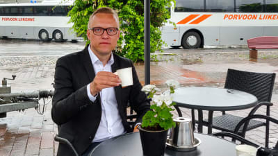 En man i kavaj sitter vid ett kafébord och håller ien pappmugg med kaffe.