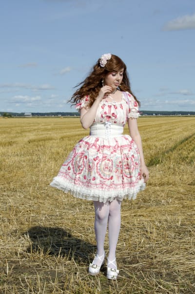 En kvinna klädd i så kallade Lolitakläder - en fluffig vit och rosa klänning med spetsar på. Hon har vita högklackade skor och blommor i håret. Hon står på en nyklippt åker.