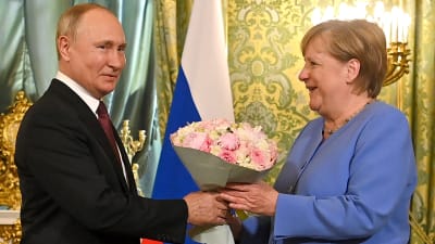 En Man i kostym överräcker en bukett med ljusröda rosor till en kvinna klädd i blått. I bakgrunden en guldfärgad vägg och en rysk flagga.