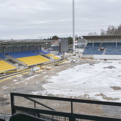 Fotbollsplanen på Kuppisstadion med snö och grävmaskiner ovanpå, i bakgrunden ser man två läktare.