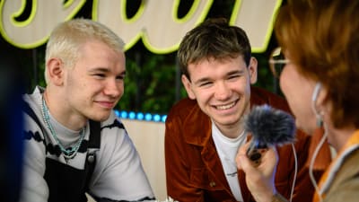 artisttvillingarna Marcus och Martinus ler mot en kvinna med kort rätt hår med bandspelare i handen som intervjuar dem för radio