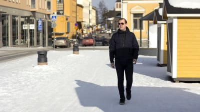 En man som går på trottoaren. Det är en solig vinterdag. Runt omkring honom finns flera gula hus. Han bär solglasögon.