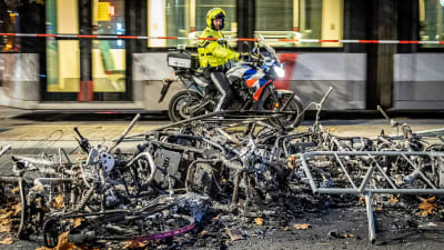 I förgrunden förkolnade cyklar och annan bråte. I bakgrunden en man i gul jacka och hjälm på en motorcykel.