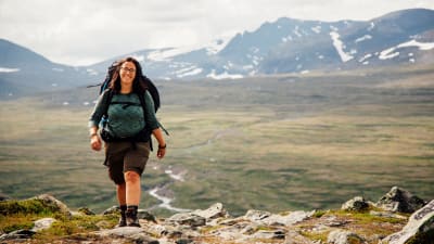 En kvinna iklädd vandringskläder och ryggsäck kommer gående över fjället. I bakgrunden syns berg.