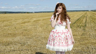 En kvinna klädd i så kallade Lolitakläder - en fluffig vit och rosa klänning med spetsar på. Hon har vita högklackade skor och blommor i håret. Hon står på en nyklippt åker.