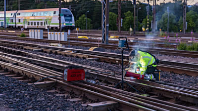 En person reparerar ett järnvägsspår. Ett tåg syns i bakgrunden.