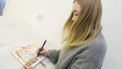 Caroline Eriksson har långt blont hår och en grå tröja. Hon lutar sig över ett bord med ett häfte framför sig. I handen håller hon en penna och hon ritar en bokhylla i häftet.