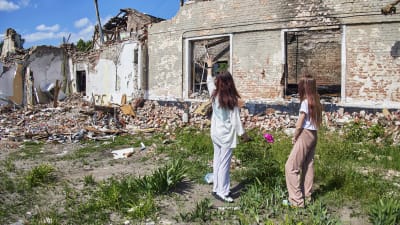 Kaksi lasta seisoo nurmella ja katsoo kohti tuhoutunutta rakennusta. Toinen lapsista on ojentanut kätensä kohti raunioita.