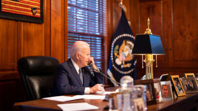 Presidentti Joe Biden puhuu lankapuhelimessa Vladimir Putinin kanssa, isolla pöydällä on perheenjäsenten valokuvia. 