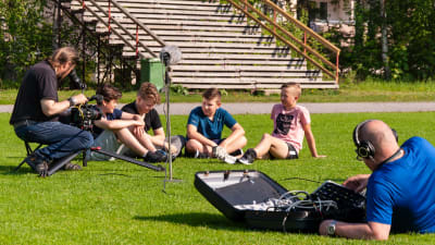 En filminspelning. Fyra pojkar sitter på en fotbollsplan, en fotograf och en ljudtekniker syns också i bild.