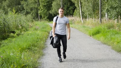 En man iklädd svarta jeans och en grå t-skjorta går på en grusväg kantad med grönt gräs och blommor. Han ler och ser glad ut.