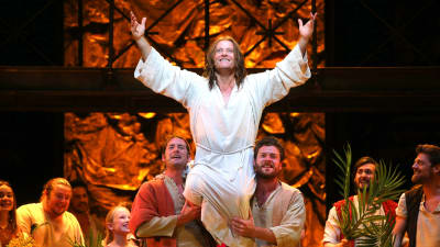 En vitlädd leende man (Jesus) som bärs upp av två män. I bakgrunden skrattande, glada människor.