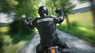 En motorcyklist i svart hjälm och svarta läderkläder sedd bakifrån, kör på en smal väg med träd och grönska på båda sidorna. Skakig bild.