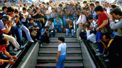 Diego Maradona är på väg upp på stadion och omringas av fotografer.