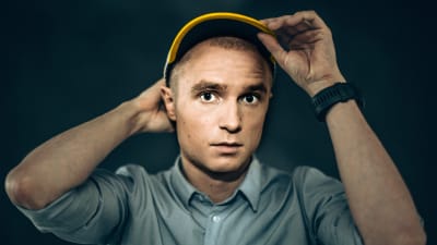 Yle Puheen Juuso Pekkinen katsoo suoraan kameraan ja pitää samalla kiinni päässän olevasta lippalakista.