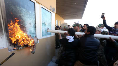 En folksamling krossar fönster på en byggnad med en stång. Ett trasigt fönster brinner.