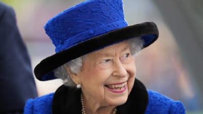 Drottning Elizabeth ler och tittar åt sidan. Hon har på sig en blå kavaj och en hatt i samma nyans. 