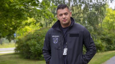 En man iklädd en jacka som det står "security" på. I bakgrunden syns träd med gröna löv. 