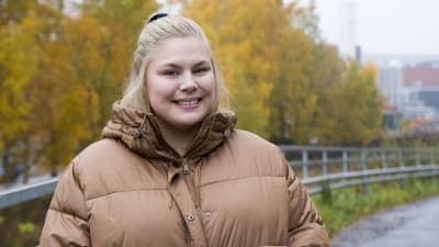 En blond kvinna iklädd en tjock beige dunjacka. Hon ler. I bakgrunden syns träd med gula höstlöv. 