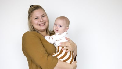 En blond kvinna iklädd en beige skjorta. Hon ler och håller i en liten baby som också ler. 