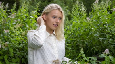 En ung kvinna med blont hår och vit skjorta står framför en häck med rosa blommor. Det är sommar.