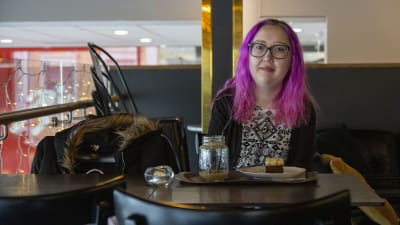 En kvinna med rosa hår. Hon sitter vid ett bord i ett kafé. Framför sig har hon en kaffe och en kakbit.