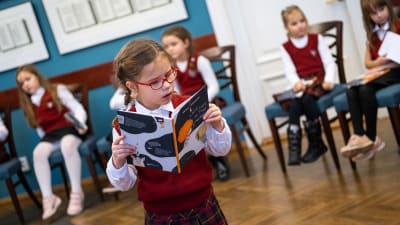 En flicka i skoluniform och röda glasögon står och läser en bok i en sal, där man ser flera andra flickor i bakgrunden som sitter på stolar.