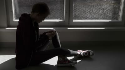 En ung person sitter på ett fönsterbräde och tittar ner på sin telefon.