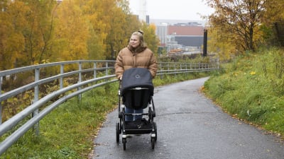 En blond kvinna iklädd en tjock beige dunjacka. Hon går på en trottoar med en barnvagn. I bakgrunden syns träd med gula höstlöv. 