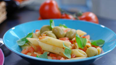 En portion penne pasta med tomatsås, garnerad med oliver och färska örter. Portionen är serverad på ett himmelsblått fat.