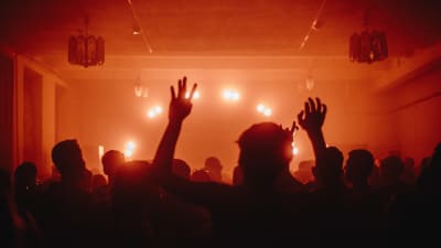 Personer dansar med ryggen mot kameran i ett rött ljus i ett dunkelt rum. Personen närmast kameran håller upp sina händer mot taket.