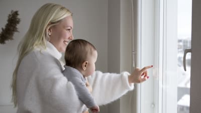 En kvinna med ett barn i famnen. Hon ler. De tittar ut genom ett fönster. 