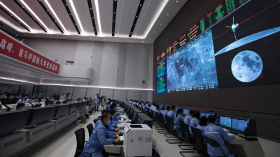 En bild tagen i kontrollrummet i Peking där många frskare sitter vid datorer och ser på en stor skärm med en bild på månen och bilder från Chang'e 5.
