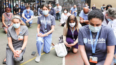 På bilden syns en stor samling sjukhusanställda som gått ner på knä för att visa sitt stöd för de antirasistiska demonstrationerna. Personerna står en bit ifrån varandra iklädda ansiktsskydd.