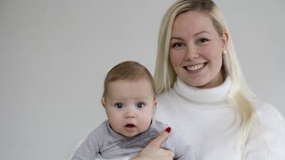 En kvinna med långt blont hår som håller en liten bebis i famnen. Båda ler. 