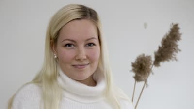 En kvinna med långt blont hår. I bakgrunden syns några torkade blommor. Hon ler. 