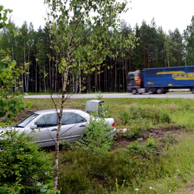 En silverfärgad bil har kört av vägen och ligger i ett dike. I bakgrunden ser man en lastbil som kör förbi.