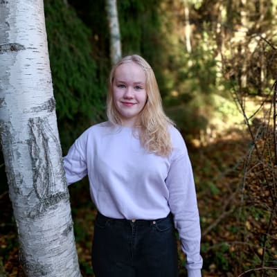 Nuori vaaleahiuksinen tyttö metsässä koivun vieressä.