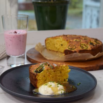 En svart fat med en portion efterrätt. På fatet finns en bit av en gul kaka med en klick creme fraiche och fikonmarmelad. I bakgrunden står en kaka, ett glas med en ljusrosa dryck och en liten skål med lingon.