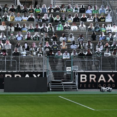 En fotbollsläktare fylld med pappfigurer i stället för riktiga människor. I förgrunden en tom fotbollsplan.