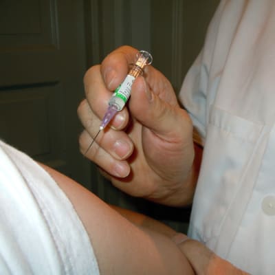 En läkare i vit rock håller i en arm, i den ena handen har hen en vaccinationsspruta.